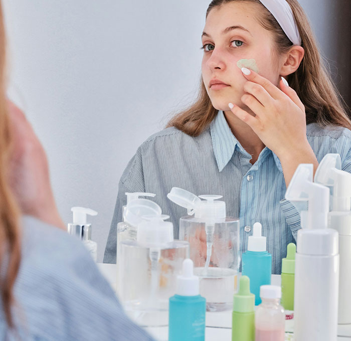 Teen using skincare products in front of a mirror, surrounded by various bottles. Teen using skincare products in front of a mirror, surrounded by various bottles.