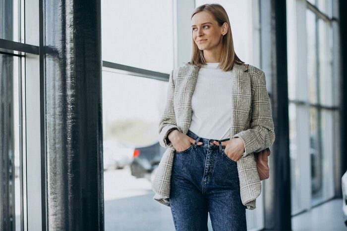 Woman standing by large windows, wearing a plaid blazer and jeans, related to insulting benefits and bonuses.