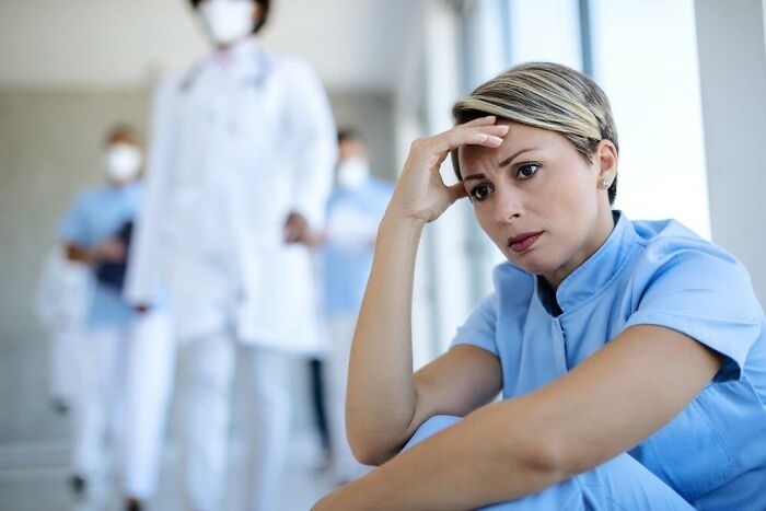 Stressed healthcare worker sitting, reflecting on insulting benefits and bonuses, with medical staff blurred in background.