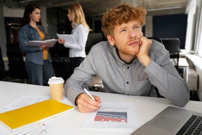 Man in an office looking thoughtful with charts, while coworkers discuss benefits and bonuses in the background.