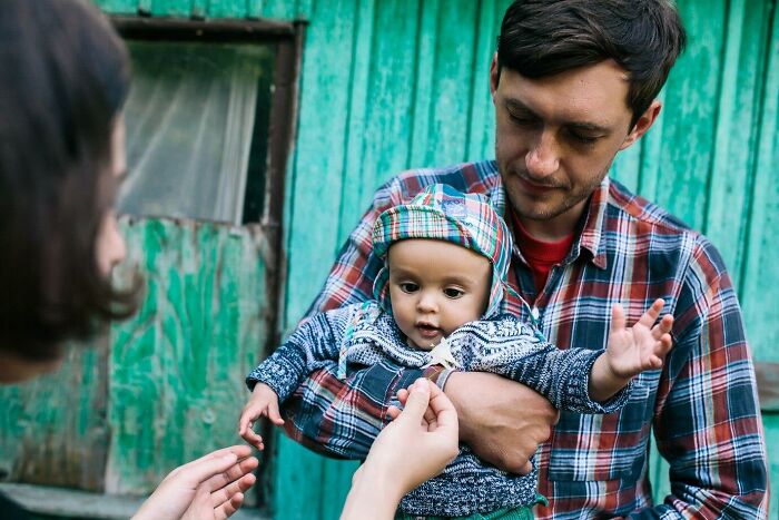 Man holding a baby, wearing plaid pattern, outside a green wood building, representing controversial takes on parenting.