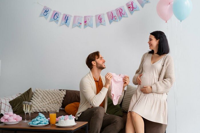 Couple at a gender reveal party, holding baby clothes, with a banner and balloons in the background.