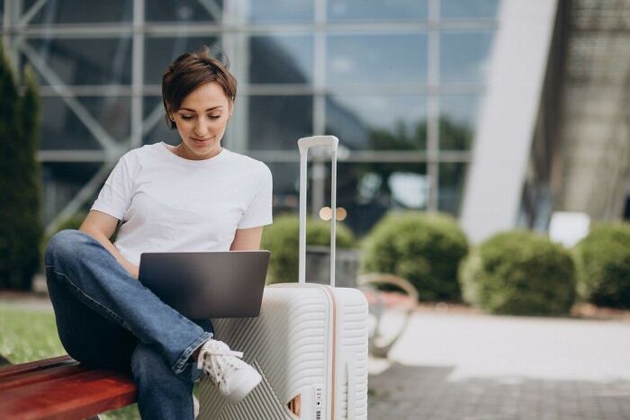 Person sitting on a bench with a suitcase, using a laptop, illustrating the romanticized idea of travel.