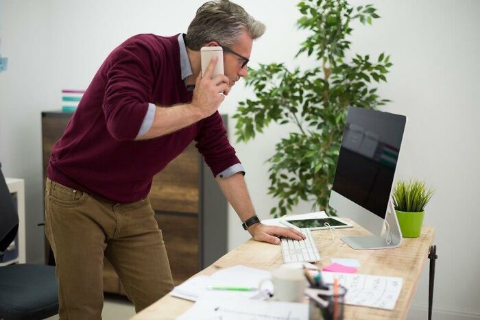 Man on phone leaning over desk with computer, depicting romanticized yet stressful work environment.