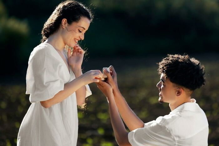Couple in a romantic engagement proposal, with the man kneeling and offering a ring outdoors.