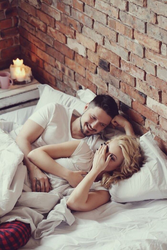 Woman and man in bed, smiling and relaxed, with warm lighting and a rustic brick wall in the background.