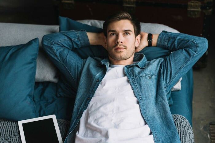 Man lying on a couch, looking thoughtful, wearing a denim shirt, with a tablet beside him.