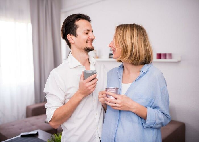 Man and woman smiling and holding coffee cups, standing in a brightly lit living room.