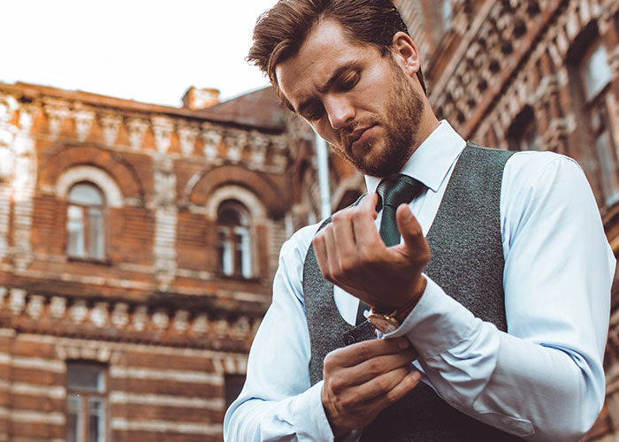 Man adjusting shirt cuffs, wearing a vest and tie, standing outside a brick building.