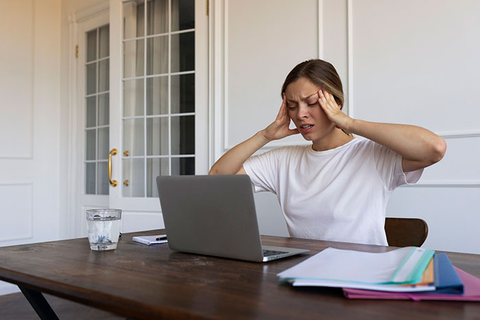 A woman sitting at a table with a laptop, appearing stressed over shut off Wi-Fi, in a room with white walls and glass doors.