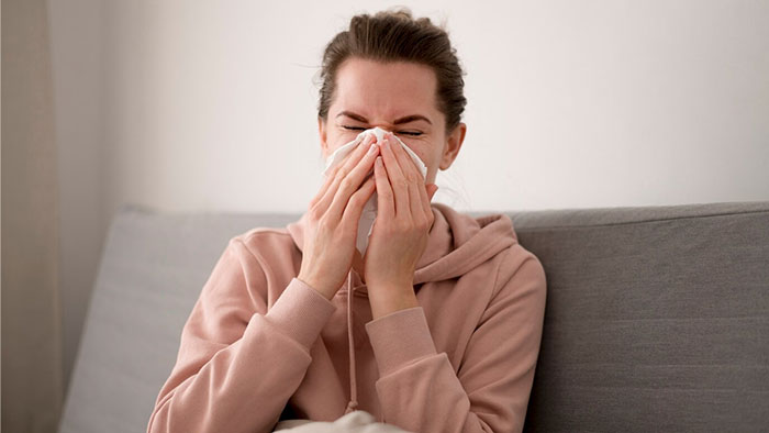 Woman sitting on a couch, sneezing into a tissue, demonstrating a crazy body life hack for dealing with colds.
