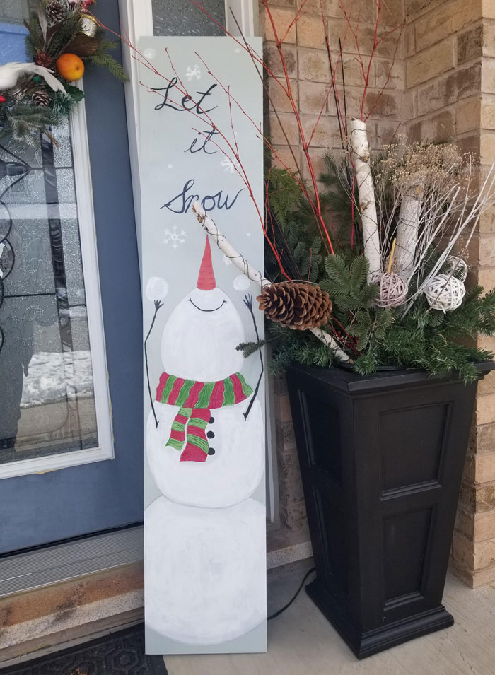 DIY Christmas decoration with painted snowman sign and festive greenery on a porch.