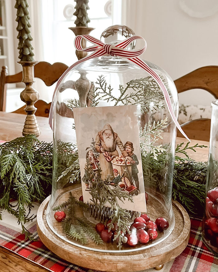 DIY Christmas decoration with vintage Santa card, greenery, and berries under a glass dome on a festive table.