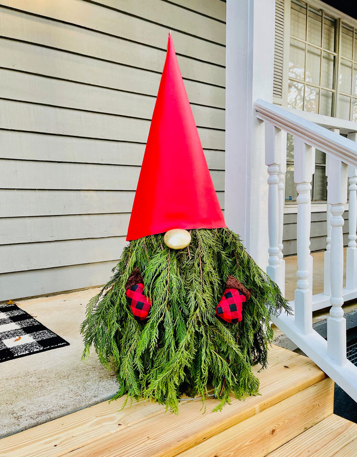 Creative DIY Christmas decoration of a gnome with pine branches and a red hat on a porch.