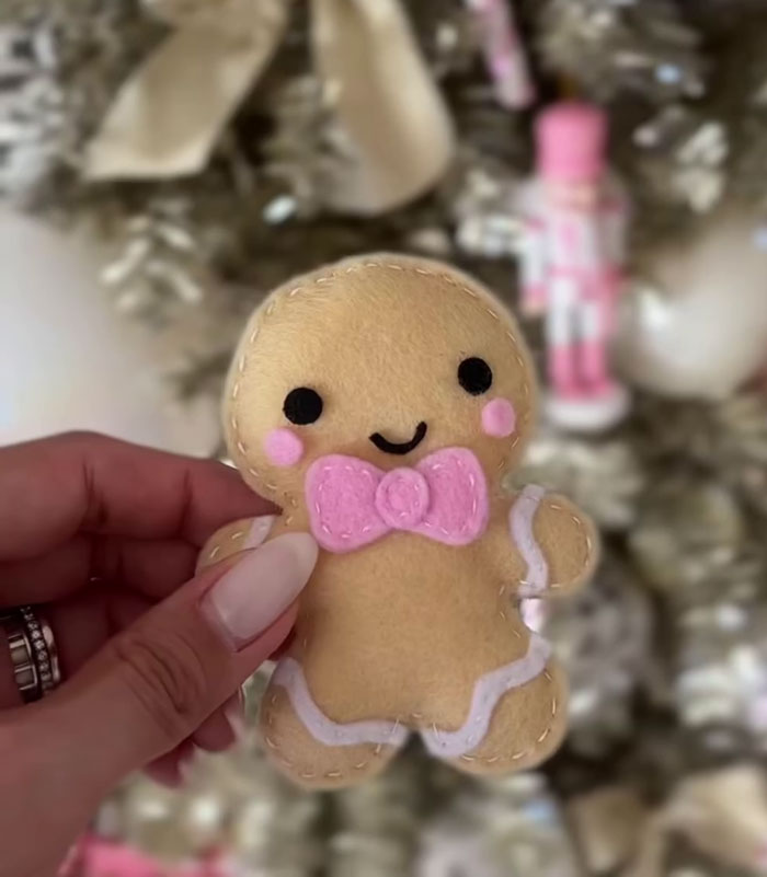 Hand holding a handmade felt gingerbread ornament with pink bow in front of decorated Christmas tree.