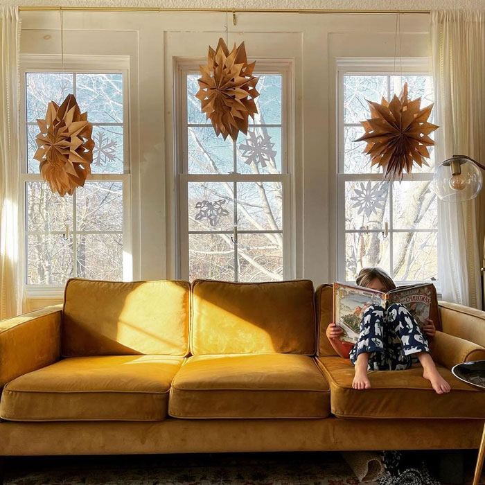 Child reading on a yellow sofa with DIY Christmas decorations hanging in front of windows.