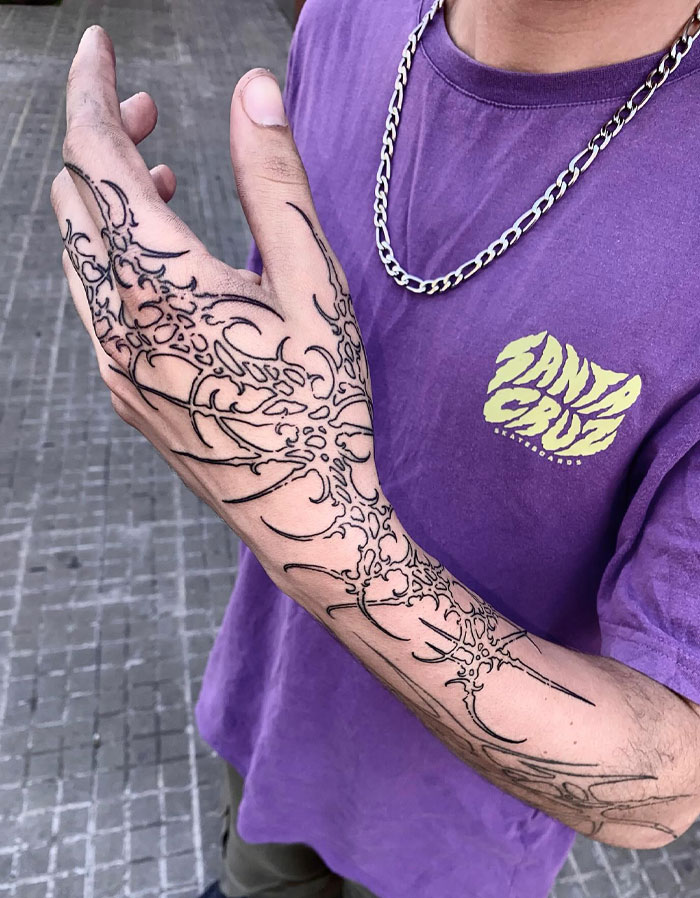 Intricate cybersigilism tattoo on a person's arm, with a purple Santa Cruz t-shirt and a silver chain necklace.