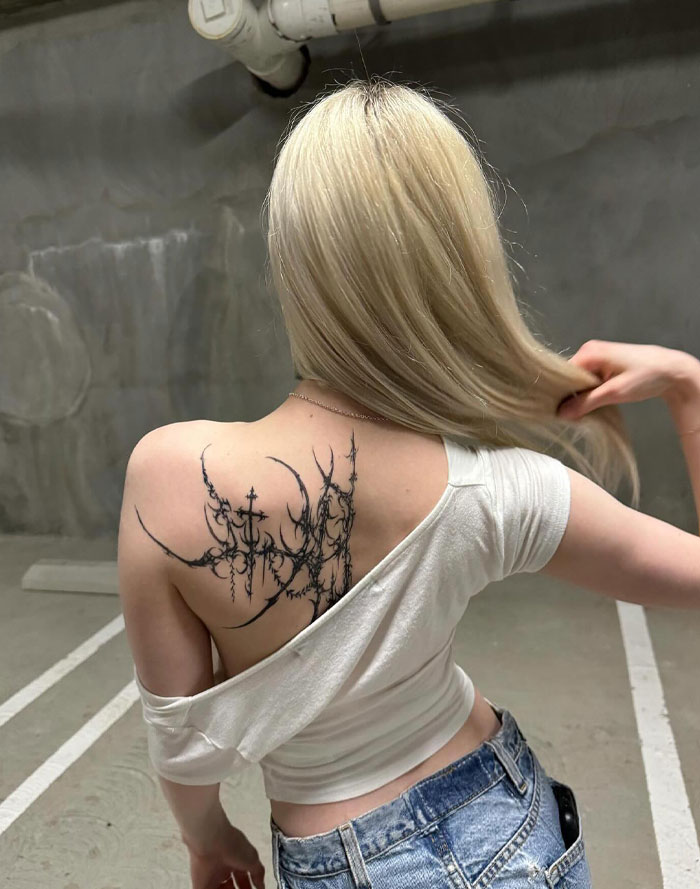 A woman with a cybersigilism tattoo on her shoulder, wearing a white top and jeans, standing in a parking garage.