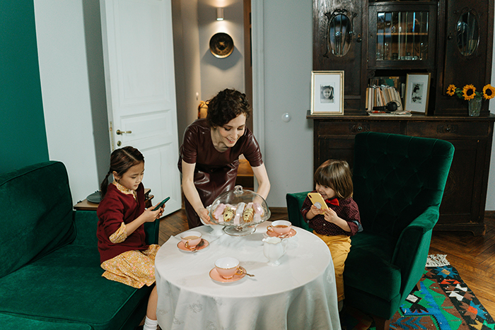A woman serves cake to two children seated at a table in a cozy living room setting, with a focus on family and home.
