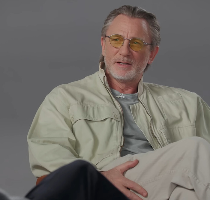 Daniel Craig in casual attire wearing yellow glasses, seated in an informal setting discussing a scene.