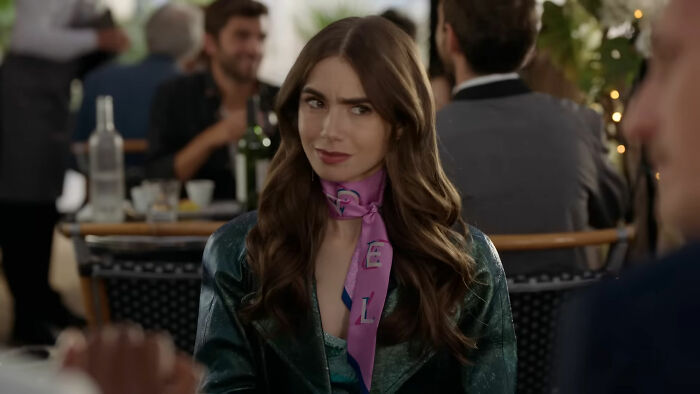 Woman smirking in a lively restaurant scene, dressed in a leather jacket and pink scarf.