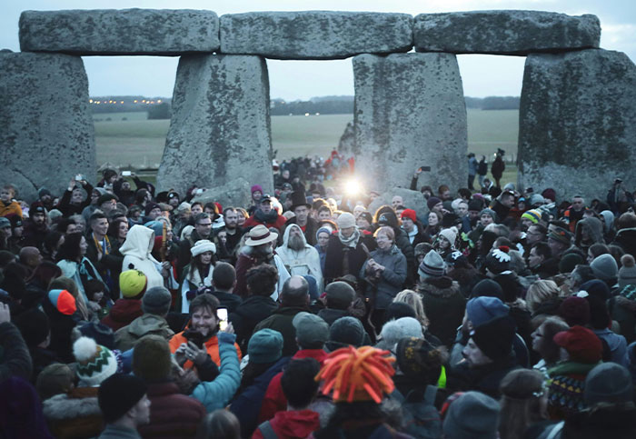 A large crowd gathers at Stonehenge, depicting a diverse group of people in winter clothing, with ancient stones in the background.