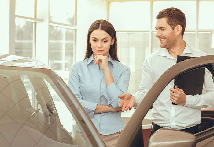 Customer question confusion; a car salesperson gestures, woman ponders beside a vehicle in a showroom.