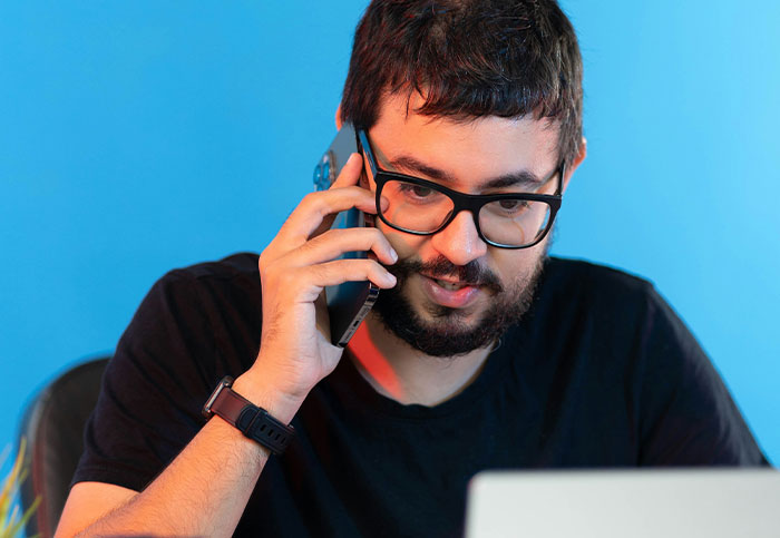 Man with glasses on phone, puzzled, experiencing a dumb customer query, in a casual setting with a blue background.
