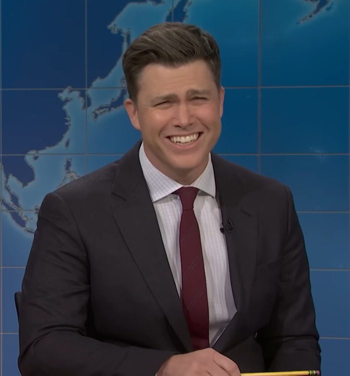 Colin Jost on set, wearing a suit and smiling, reading jokes about Scarlett Johansson.