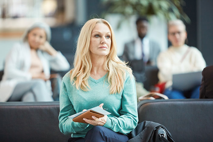 Woman with blonde hair sitting in an airport waiting area, holding a tablet, surrounded by other travelers. Woman with blonde hair sitting in an airport waiting area, holding a tablet, surrounded by other travelers.