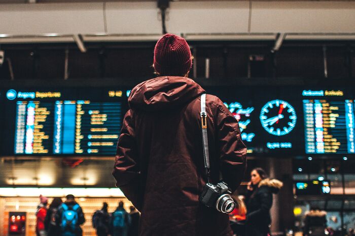 Person in a coat and beanie with a camera, looking at a flight timetable.