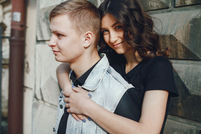 Young woman hugging a young man from behind in an outdoor setting, both smiling.