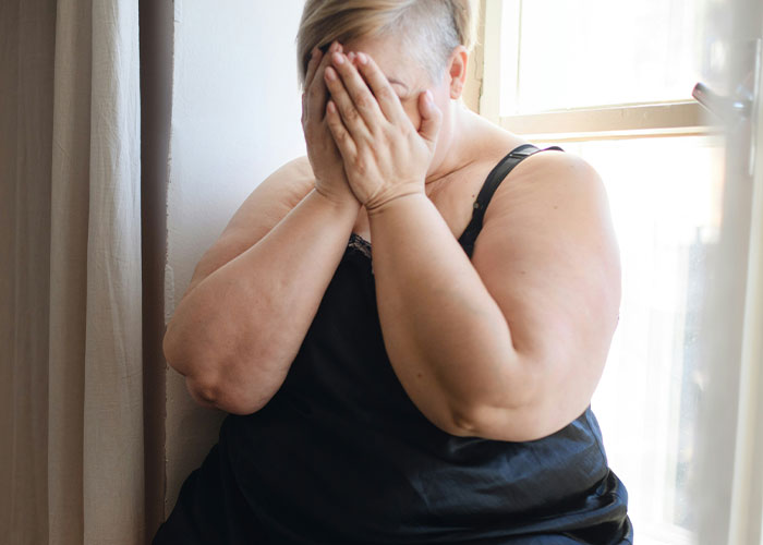 Person in black camisole sitting by a window, covering face, expressing distress about fat-shaming. Person in black camisole sitting by a window, covering face, expressing distress about fat-shaming.
