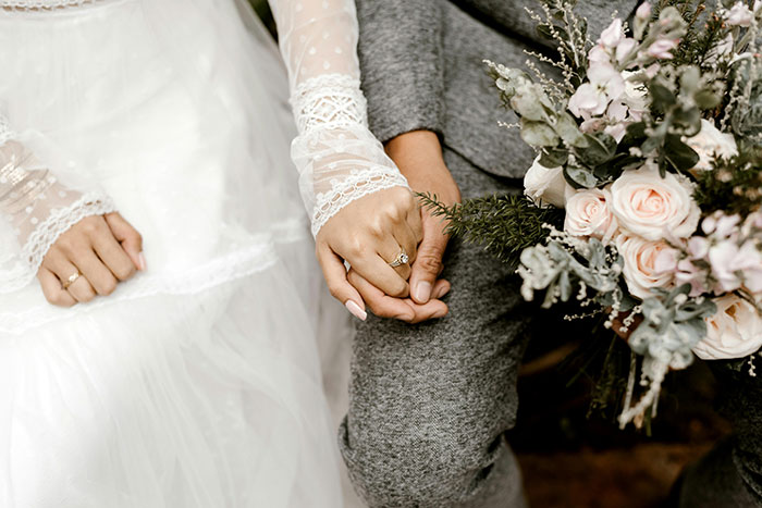 A couple holding hands, dressed in wedding attire, with a bouquet of flowers.
