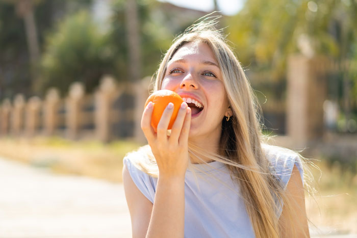 Woman joyfully eating an orange, enjoying fruit in a sunny outdoor setting.