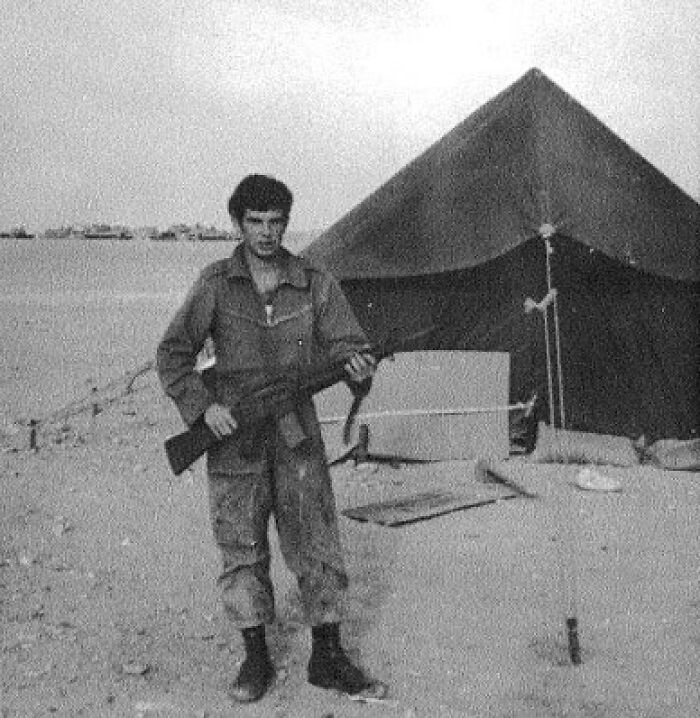 Young soldier with a rifle stands in front of a military tent in 1973, illustrating a scene from that era.