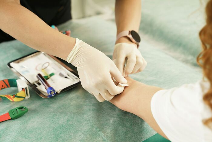Medical professional drawing blood, wearing gloves and a smartwatch, with medical tools nearby on a green surface.