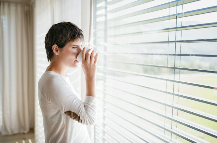 Person drinking coffee, gazing out window blinds, representing stories of nice guys.
