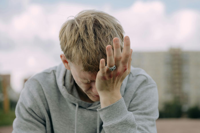 Man in gray hoodie looking regretful with hand on face, wearing engagement ring.