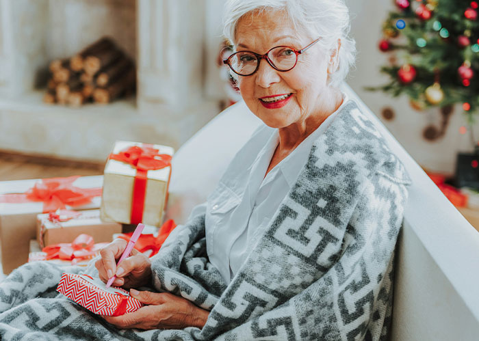 Elderly woman writing on a gift tag, surrounded by wrapped gifts in a festive setting.