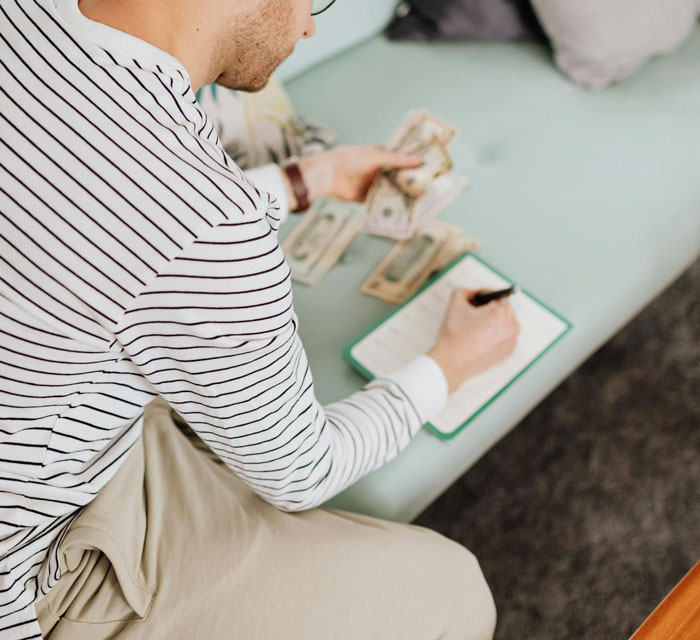 Man in striped shirt counting money, writing in notebook, highlighting cost concerns for a destination wedding. Man in striped shirt counting money, writing in notebook, highlighting cost concerns for a destination wedding.