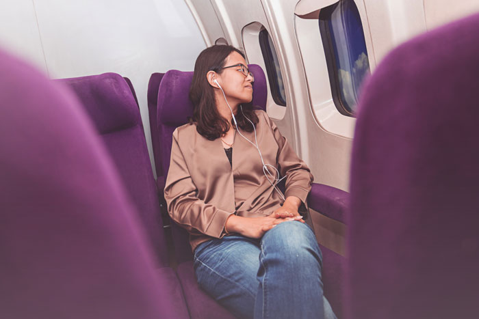 Tall woman relaxing comfortably in an airplane seat, wearing headphones and a beige jacket, enjoying her window view.