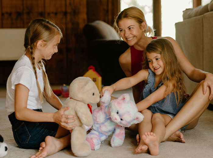 Mother and daughters playing with stuffed animals in a cozy living room.