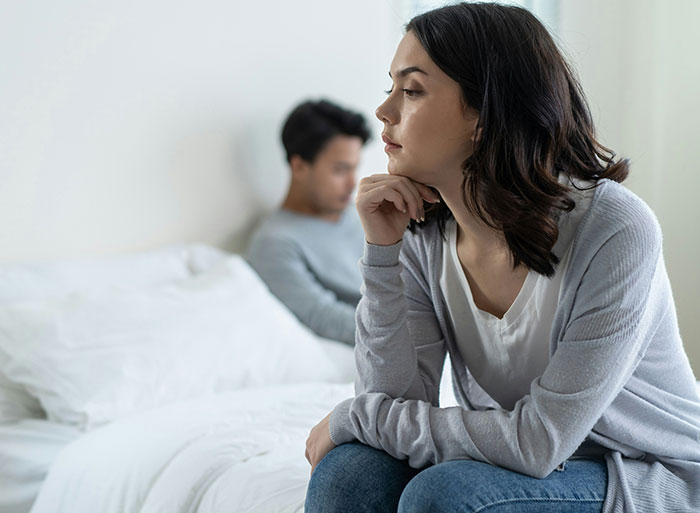 Woman deep in thought sitting on a bed, man in the background, focusing on family conflict theme.