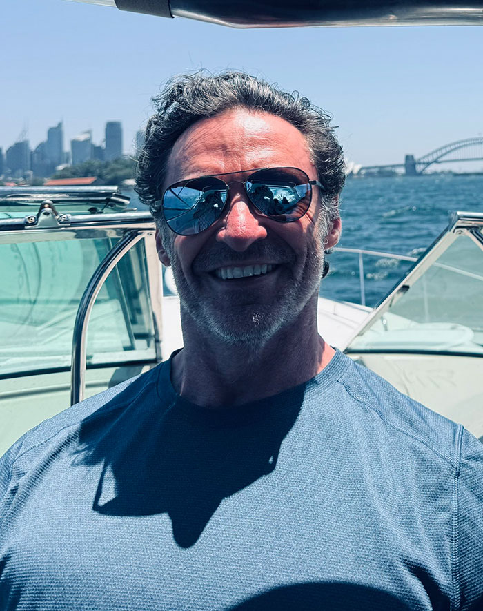 Man in sunglasses and blue shirt on a boat, with a city skyline and bridge in the background, sparking rumors. Man in sunglasses and blue shirt on a boat, with a city skyline and bridge in the background, sparking rumors.