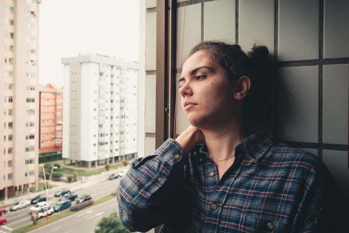 Woman looking frustrated by a window, wearing a plaid shirt, contemplating after an interruption during her job test.