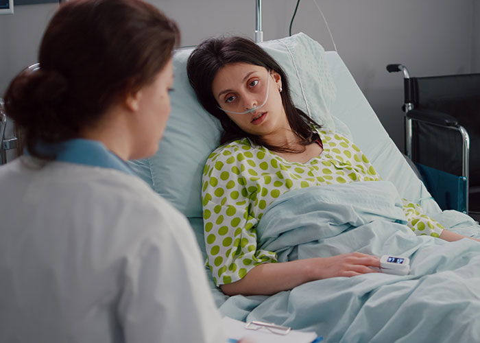 Woman in hospital bed with doctor, discussing surgery while using an oxygen tube, highlighting a husband's defensive absence.