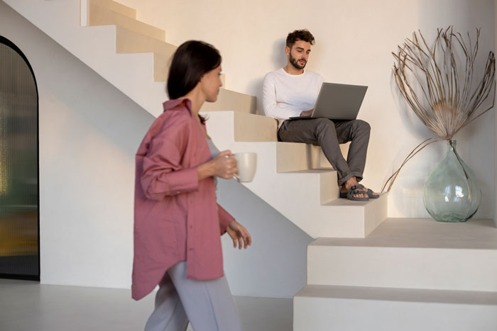 Man sitting on stairs with laptop as woman walks by holding a mug