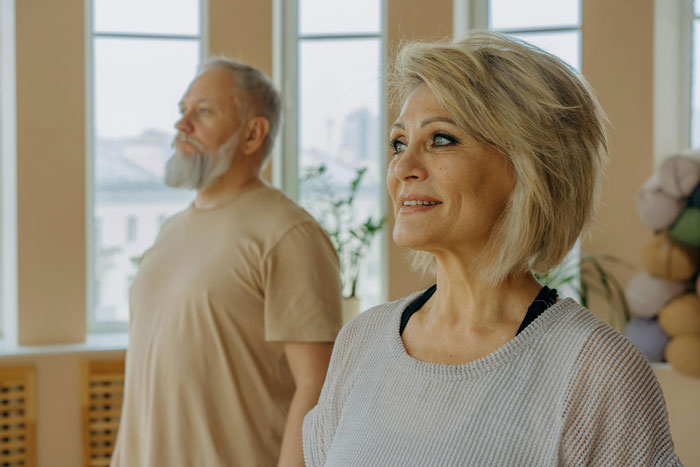 Older couple standing indoors with relaxed expressions, practicing mindfulness in a bright room.