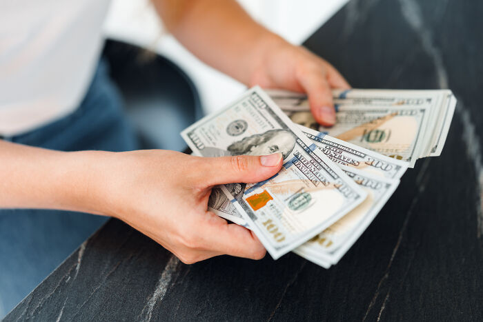 Hands holding several hundred-dollar bills on a dark marble table.